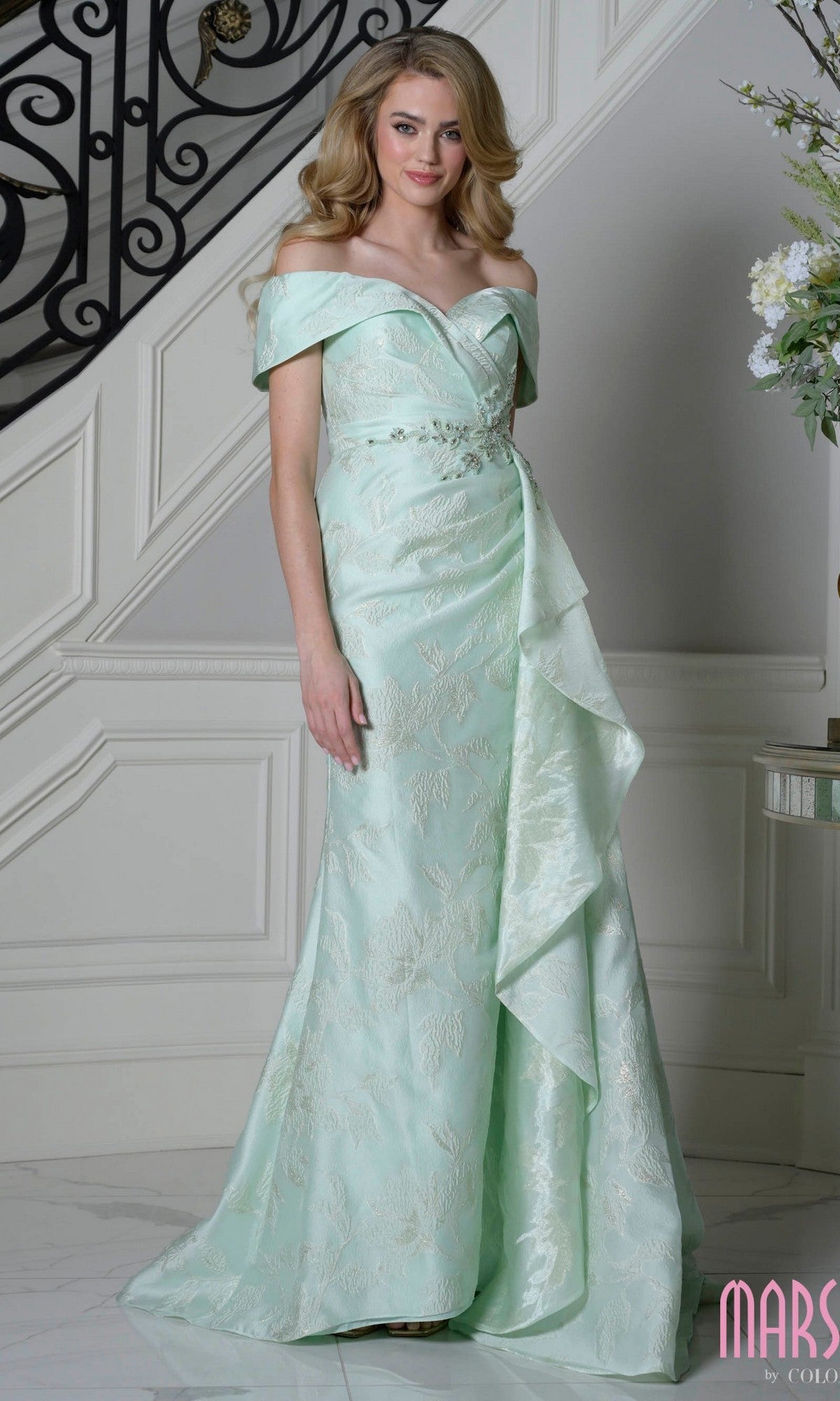 A woman stands indoors near a staircase wearing the Marsoni MV1401 long formal dress in mint. This elegant off-the-shoulder gown features floral patterns, ruffled details, and beautiful textured jacquard fabric for added charm.